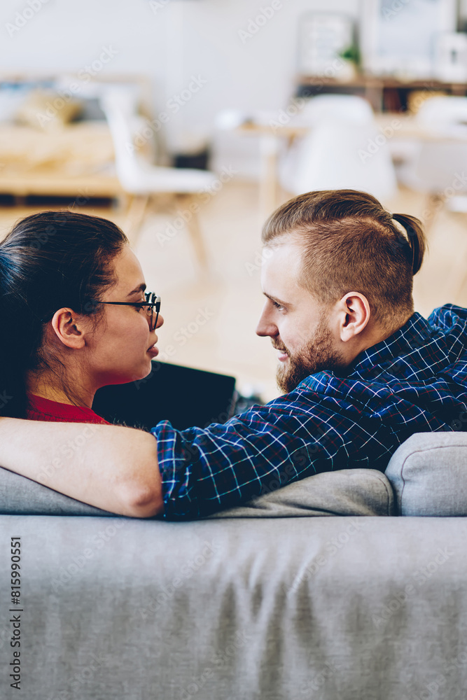 Back view of romantic couple sitting on comfortable couch in living room of apartment,hipster guy in love looking at her girlfriend next to him on sofa using laptop at contemporary home interior Back view of romantic couple sitting on comfortable couch in living room of apartment,hipster guy in love looking at her girlfriend next to him on sofa using laptop at contemporary home interior