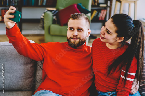 Young romantic marriage posing for common picture on smartphone camera sitting on couch at home interior,hipster couple in love making selfie on modern mobile phone for share in social networks photo