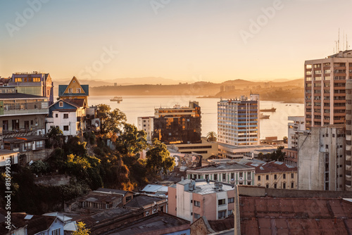 Panoramic view of the city of Valparaiso, Chile, featuring traditional houses built on hillsides, colorful murals, modern towers and a bay with ships at anchor.