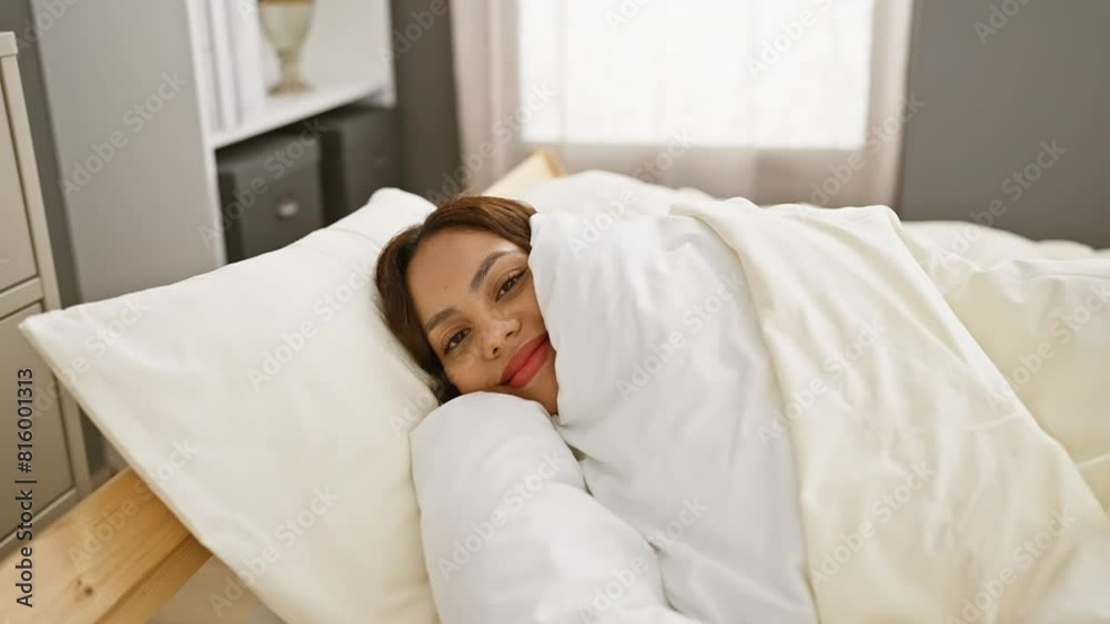 Young woman wrapped in a white duvet smiles softly while resting in a bedroom with a modern decor. Young woman wrapped in a white duvet smiles softly while resting in a bedroom with a modern decor.