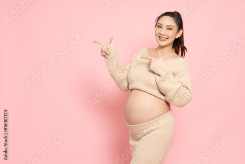 Happy Asian Pregnant woman standing and pointing to copy space isolated on pink background, Motherhood and Pregnancy Concept