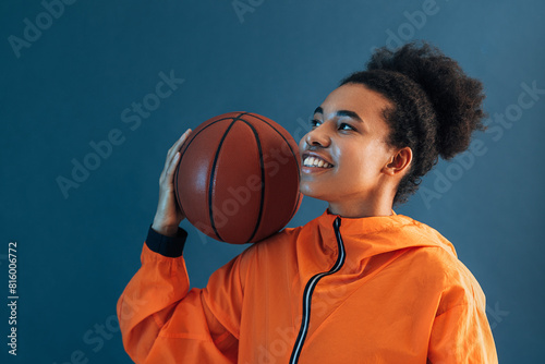 Smiling woman in orange sports clothes holding a basketball on her shoulder. Young female basketball player posing over blue backdrop.