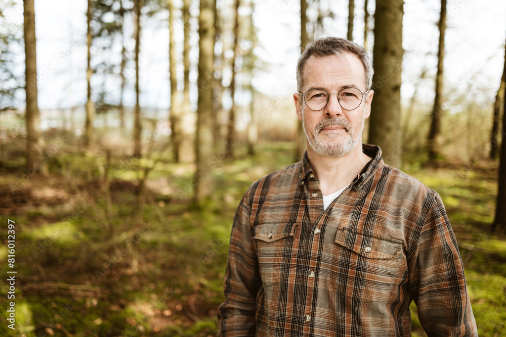 Portrait of a middle-aged man standing in a serene forest wearing a plaid shirt and glasses, exuding confidence and tranquility. Portrait of a middle-aged man standing in a serene forest wearing a plaid shirt and glasses, exuding confidence and tranquility.