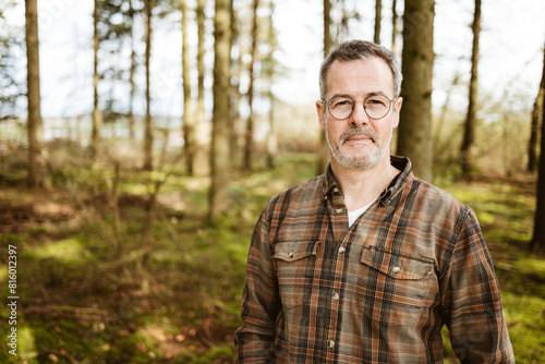 Portrait of a middle-aged man standing in a serene forest wearing a plaid shirt and glasses, exuding confidence and tranquility.