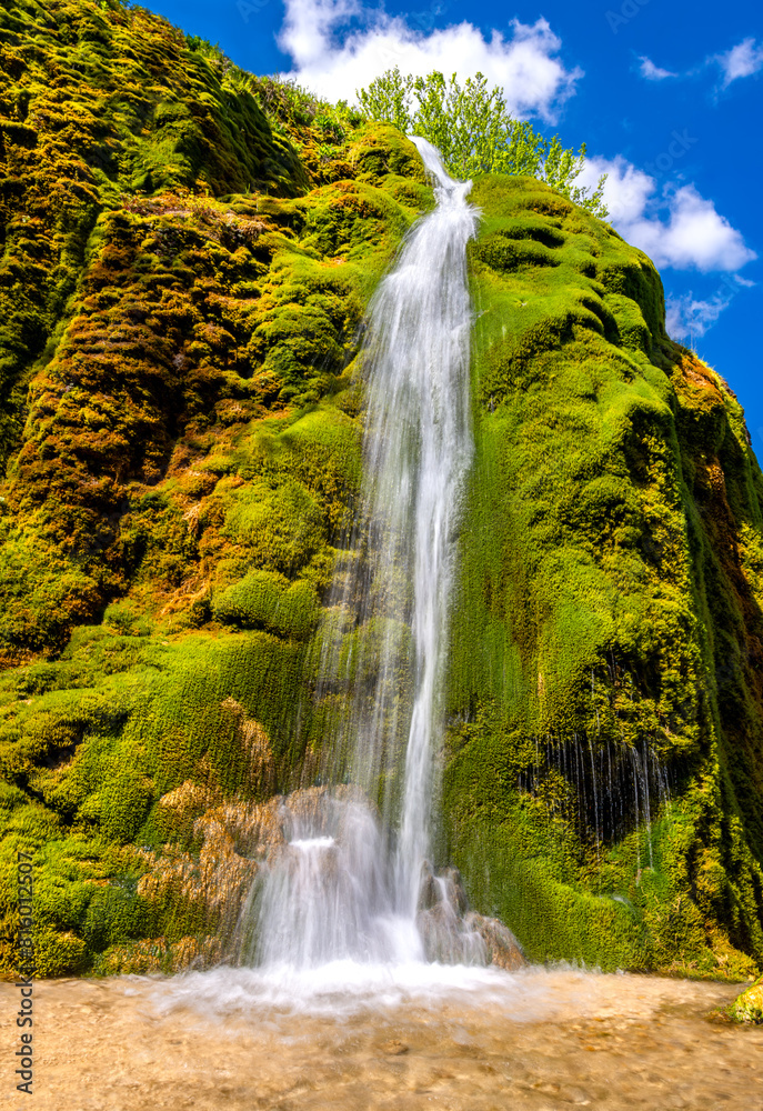 Fototapeta premium The “Dreimühlen Wasserfall“ formed from limestone deposits at the Ahbach creek in the Eifel, Germany. Long exposure of colorful tall cascade on a sunny May day. Tourist attraction and natural reserve.