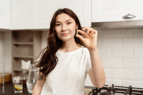 Confident Young Woman In Kitchen Showing Omega-3  Modern Interior Background. Concept Of Satisfaction  Approval  Good Service. Natural Light  Casual Style.