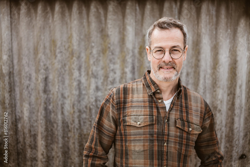 Smiling mature man with glasses and a plaid shirt standing outdoors against a textured wall, exuding confidence and warmth.