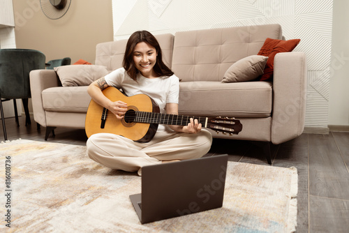 Joyful Young Woman Playing Guitar At Home, Sitting On Floor With Laptop Nearby, Expressing Creativity And Relaxation In Comfortable Modern Living Room.