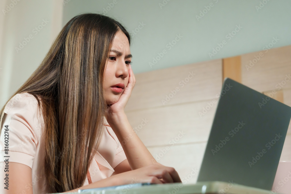 Young Asian woman with thoughtful expression using laptop in bedroom. Wearing light pink blouse, long hair. Hand on face, concentrated. Modern, comfortable bedroom, working from home.