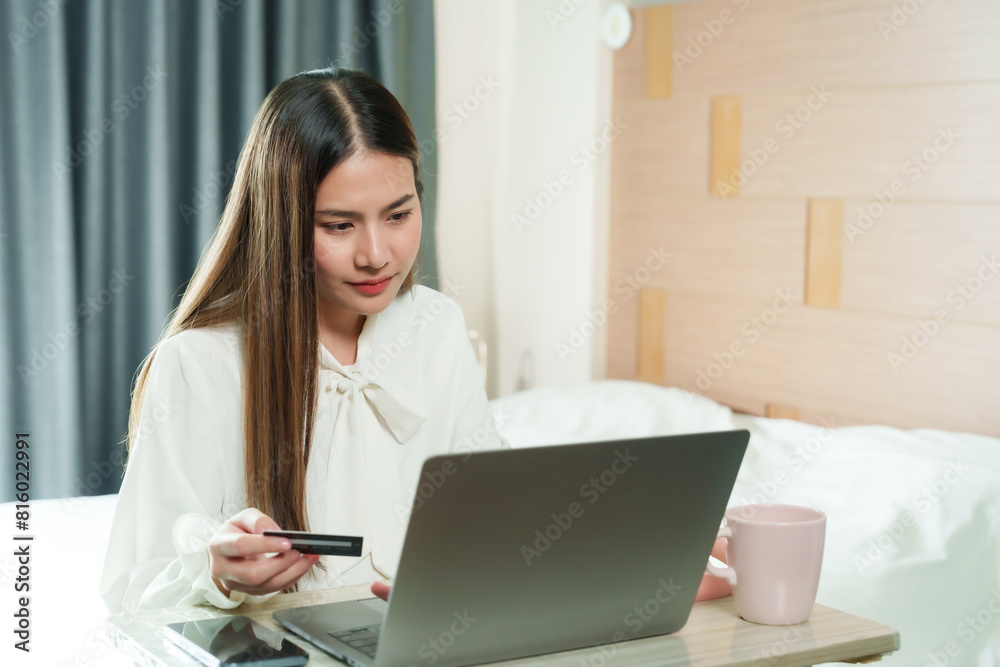 Young Asian woman holding credit card, looking at laptop screen, serious expression, making online payment in cozy room. Light wood background, pink mug on desk, smartphone beside her. Young Asian woman holding credit card, looking at laptop screen, serious expression, making online payment in cozy room. Light wood background, pink mug on desk, smartphone beside her.