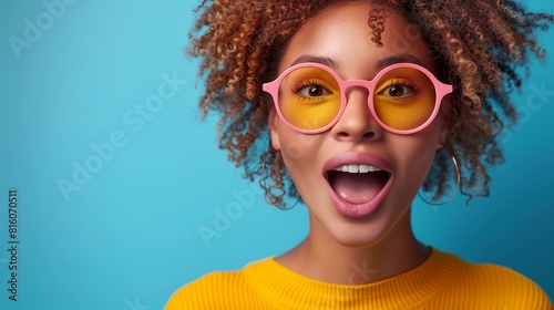 Influencer culture  young woman curly hair wearing yellow sweater pink sunglasses surprised expression on face in front of blue background