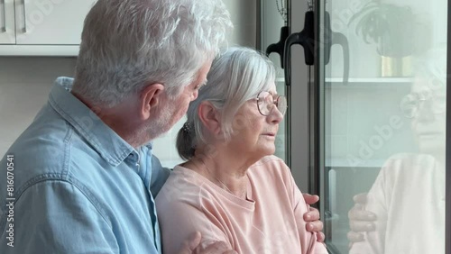 A sad elderly woman looks out the window expressing melancholy while her husband consoles her, an elderly married couple hugs and supports each other in a difficult time