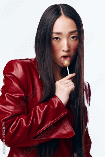 Stylish Asian Woman in Red Leather Jacket Holding Lollipop in Mouth Outdoors in Urban Setting