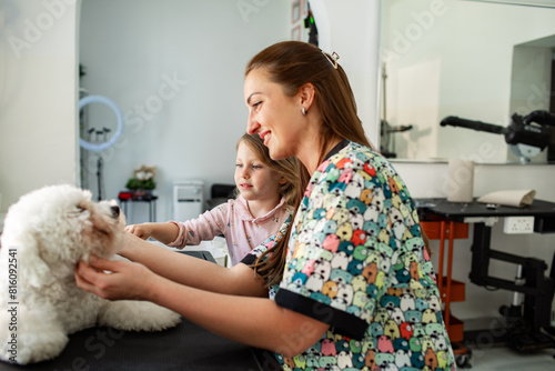 Pet groomer brushing white dog with little girl watching at salon photo