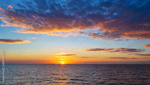 Sunset casts warm red glow over calm sea, with light breeze gently rippling surface. Fluffy clouds drift across sky, edges illuminated by setting sun's radiant light. Serene, tranquil atmosphere.