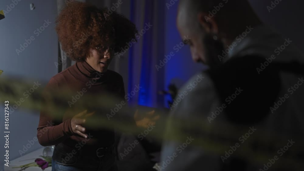 A woman explains to a man in a dimly lit, modern living room, evoking a sense of dialogue or investigation.