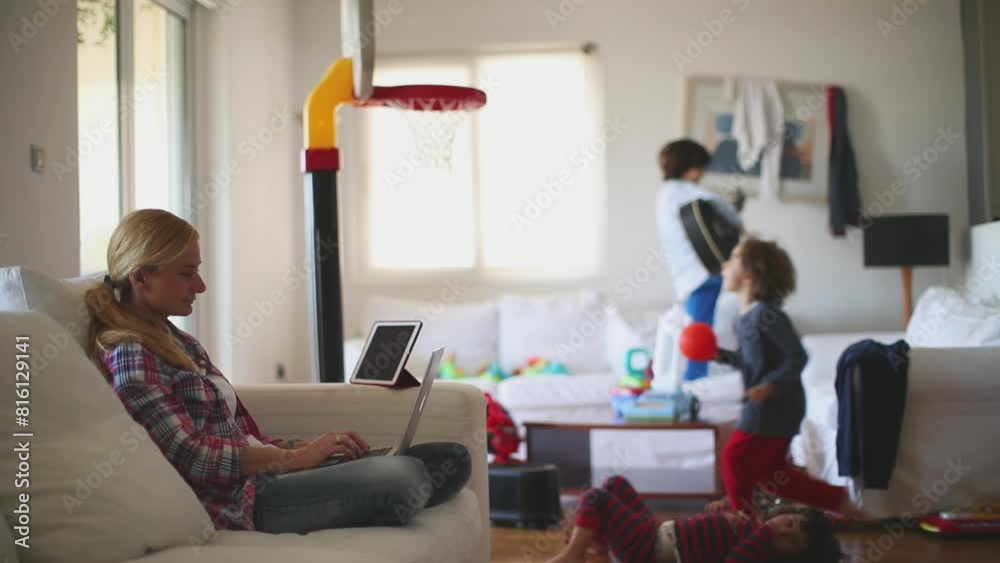 A mother works from her laptop on a couch, while her children play energetically in the living room filled with toys. A mother works from her laptop on a couch, while her children play energetically in the living room filled with toys.