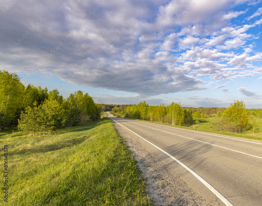Asphalt road through a field, Evening landscape. Blue sky with clouds above the horizon. early spring in the countryside, soft sunlight on the grass. Asphalt road through a field, Evening landscape. Blue sky with clouds above the horizon. early spring in the countryside, soft sunlight on the grass.