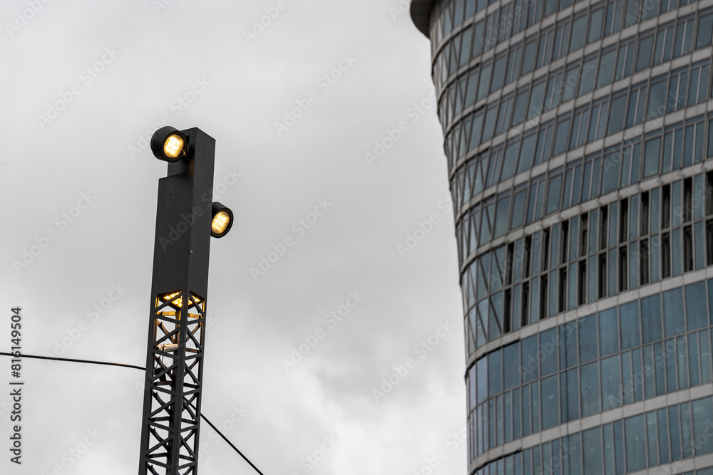 Obraz premium A modern streetlight with illuminated light bulbs stands against the backdrop of a towering skyscraper with a reflective glass facade, under an overcast sky in the city center.