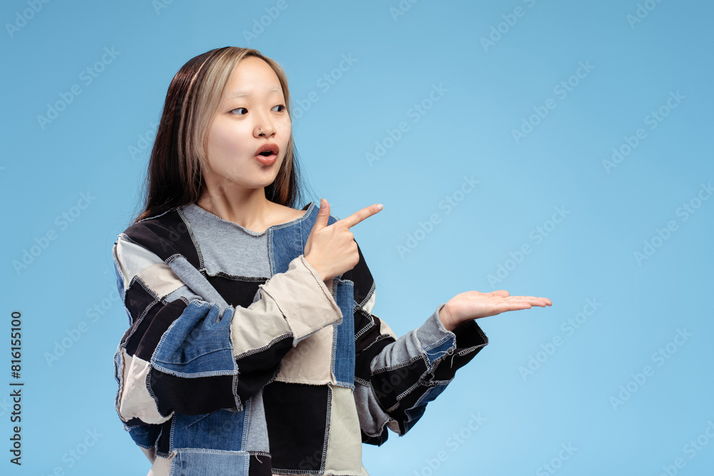 Surprised Asian girl showing by finger to copy space, looking away, isolated on blue Surprised Asian girl showing by finger to copy space, looking away, isolated on blue