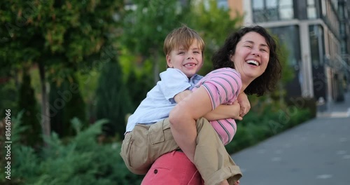 Mother taking her child on a sholders or on the back. Bright summer day in the urban background. Young caucasian mother and son kid having fun together outdoors