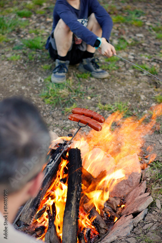 Father and son roasting sausages skewered on a grill fork over an open campfire in the evening. Grilling sausages for dinner at camping or in the garden. Summer camping in the countryside.