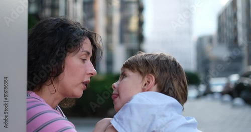 Cute caucasian brunette mother and kid son sitting outdoors face to face talking to each other. Motherhood or family day concept. 8 years old boy with mother in bright clothes enjoying summer day