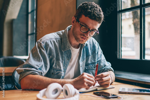 Serious male writer sitting at desktop with textbook for education and holding pen in hand while thinking on information for article, pensive hipster guy in eyeglasses enjoying autodidact indoors photo