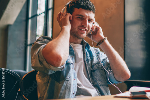 Portrait of successful hipster guy in electronic earphones looking at camera and enjoying music hobby indoors, happy smiling male meloman listening positive playlist via headphones on leisure photo