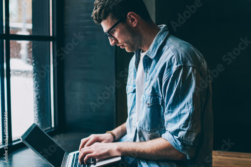 Caucasian male freelancer sitting near cafeteria window and typing new article in modern laptop application, young man blogger browsing wireless internet and updating profile in social networks