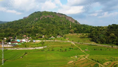 Panorama of the Nglanggeran Ancient Volcano ecotourism area in the afternoon using a drone. Aerial photo of an ancient stone hill covered with lush trees and surrounded by rice fields and countryside photo