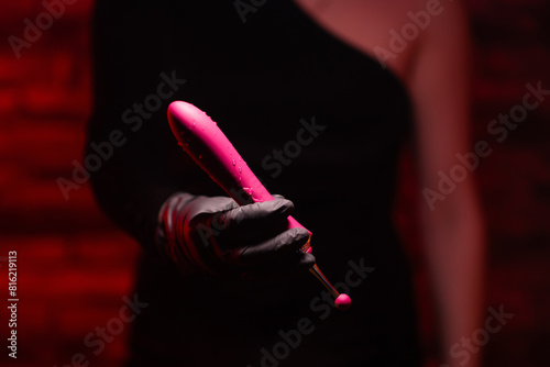 Burgundy stylish dildo for masturbation in the hand of a girl wearing black gloves on a red background. Nozzle for clitoral stimulation. Sex shop Adult store