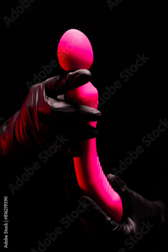 Pink stylish vibrator for masturbation in the hands of a girl in black gloves on a black background. Massager with multiple speeds. Products for sex shop, gifts for adults