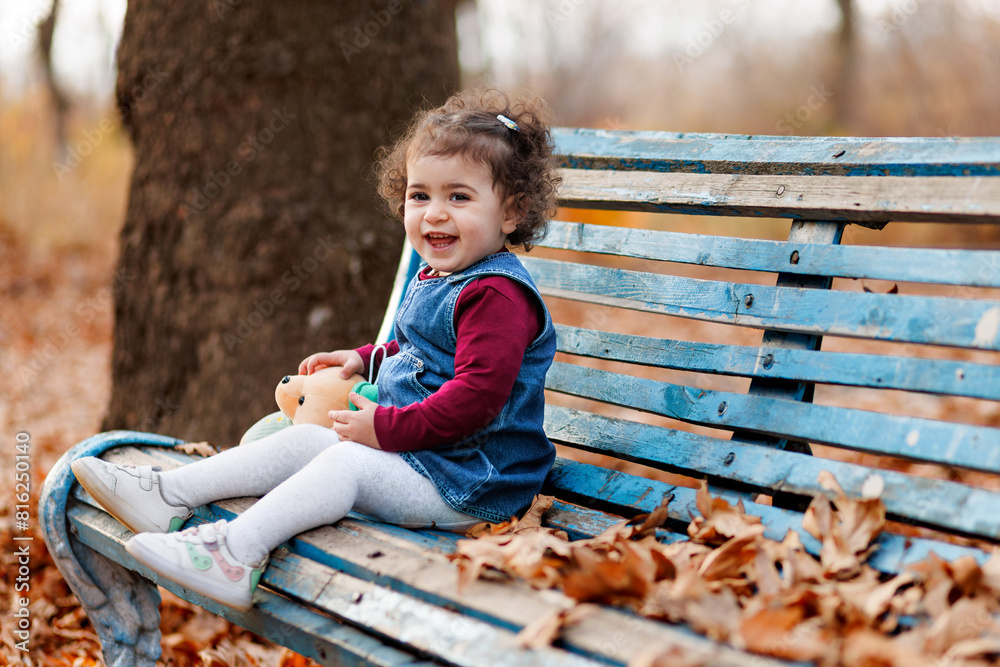 Cute laughing little girl holding her teddy bear and sitting on wooden bench amidst fallen leaves
