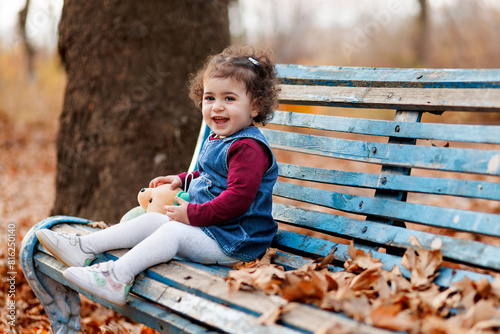 Obraz na płótnie Cute laughing little girl holding her teddy bear and sitting on wooden bench amidst fallen leaves w salonie