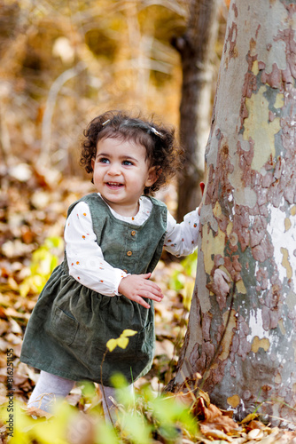 Smiling little girl standing by a tree, touching its bark, leaves at their feet