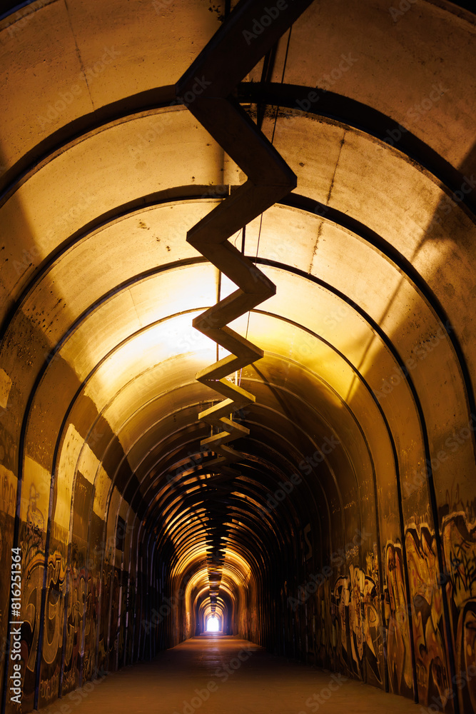 Urban, graffiti-covered tunnel with zigzag installation and warm lights leading to bright exit