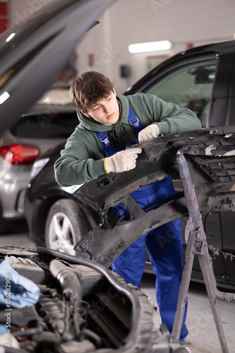 Young guy mechanic in uniform inspects car bumper in car service station