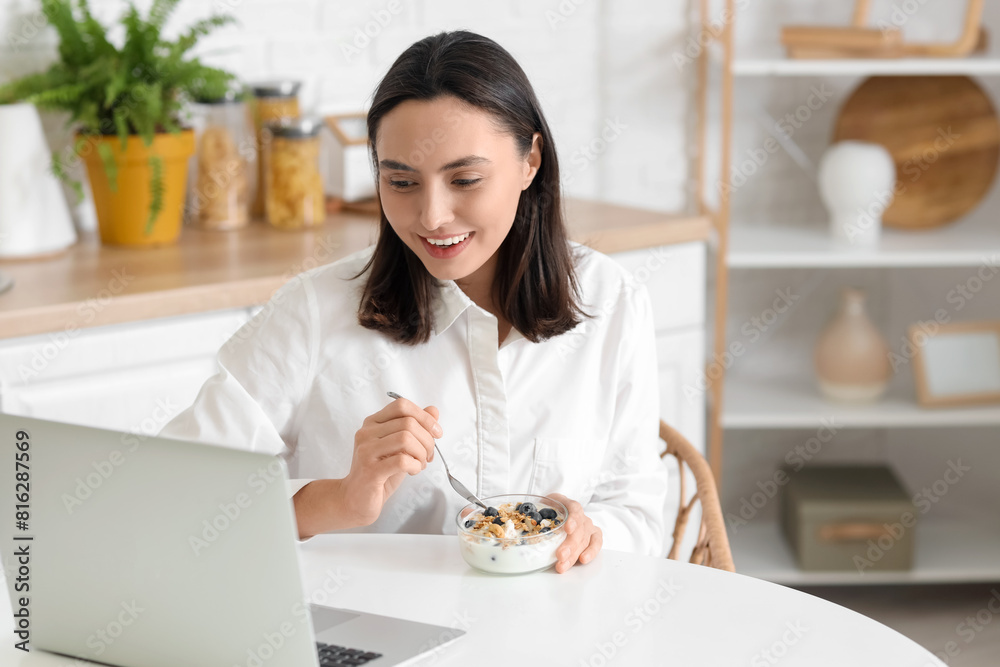 Happy young woman with bowl of tasty yoghurt and laptop at table in kitchen