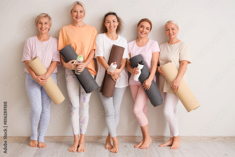 Group of mature women with yoga mats and water bottles near light wall