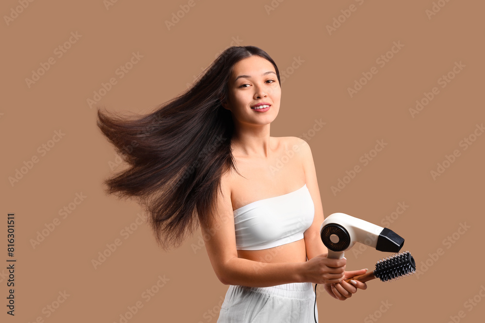 Beautiful young happy Asian woman with hairdryer and brush on brown background Beautiful young happy Asian woman with hairdryer and brush on brown background