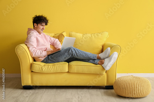 Young man resting on soft sofa with laptop near yellow wall photo