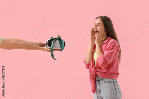 Happy young woman receiving gift VR glasses on pink background