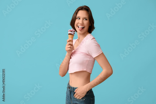Beautiful young woman with sweet ice-cream in waffle cone showing tongue on blue background