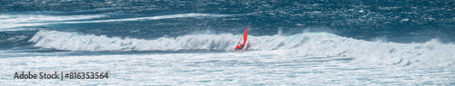 Man on a wind surfer with red sail crashing on a wave on the Pacific Ocean off the coast of Maui at Hookipa Beach, Hawaii 