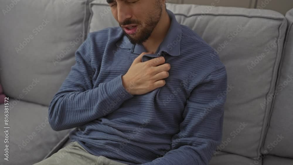 A handsome young man with a beard, feeling unwell, sits on a sofa indoors, clutching his throat. A handsome young man with a beard, feeling unwell, sits on a sofa indoors, clutching his throat.