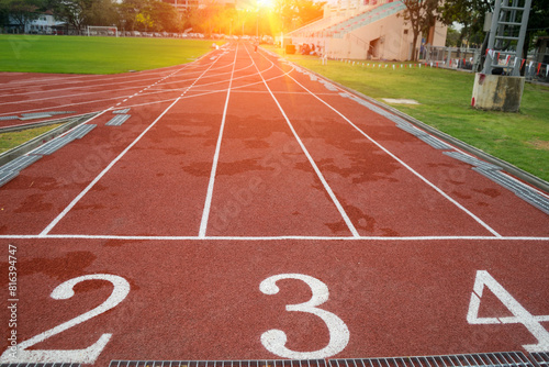Red running track in outdoor stadium with red asphalt or rubber and white markings for fitness or competition with green grass field with sunlight background.
