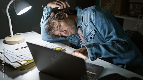 An overworked man sitting tiredly at his desk indoors, with a laptop, paperwork, and desk lamp in a dark office. photo