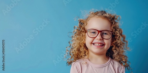 a young girl with curly hair and brown and red glasses smiles while wearing a pink shirt, standing in front of a blue wall her blue eyes and open mouth are visible, as well photo