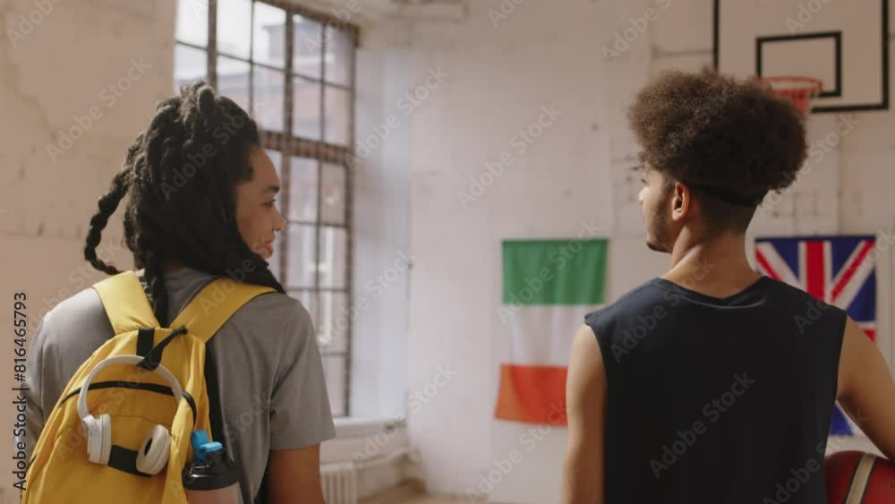 Tracking back-view slowmo of young Biracial couple of basketball players talking while walking along indoor court, arriving on training Tracking back-view slowmo of young Biracial couple of basketball players talking while walking along indoor court, arriving on training
